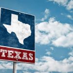 Vintage-style Texas road sign with a map outline against a blue sky with clouds.